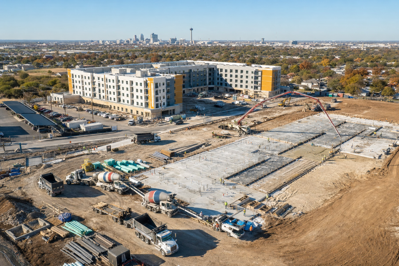 Aerial view of a large multifamily apartment construction site in San Antonio showing concrete foundation work in progress, dump trucks, ready-mix trucks, and organized staging areas during fall construction season